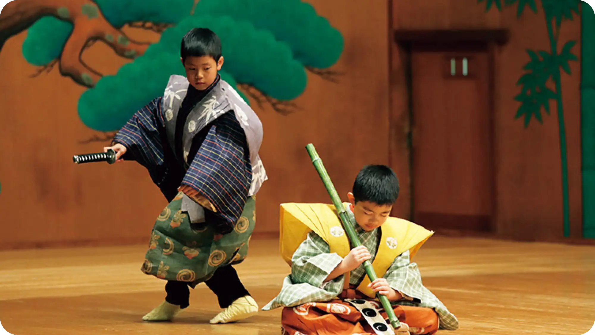 竹を持って座っている男の子と、その後ろで刀を手に立っている男の子